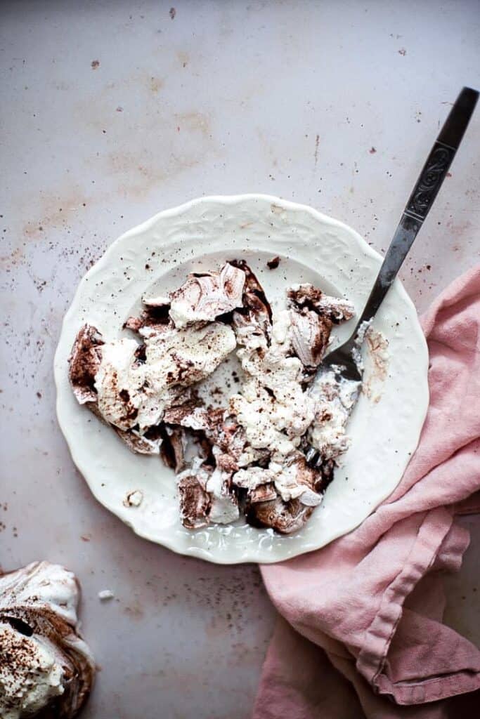A squashed chocolate swirl meringue, on a white plate, with whipped cream and a pink linen napkin.